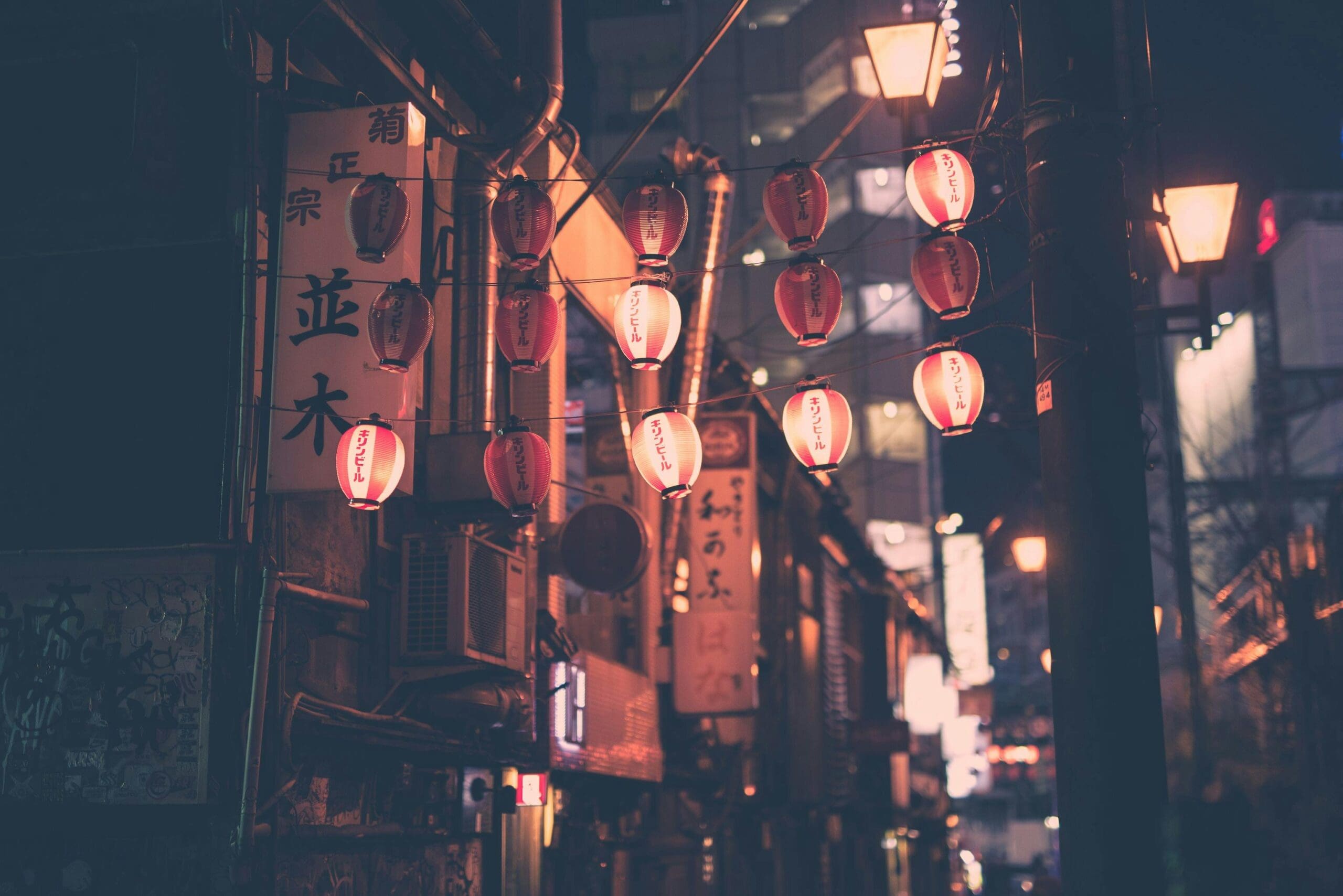 Red paper lanterns hanging above a narrow street in a Japanese city at night.