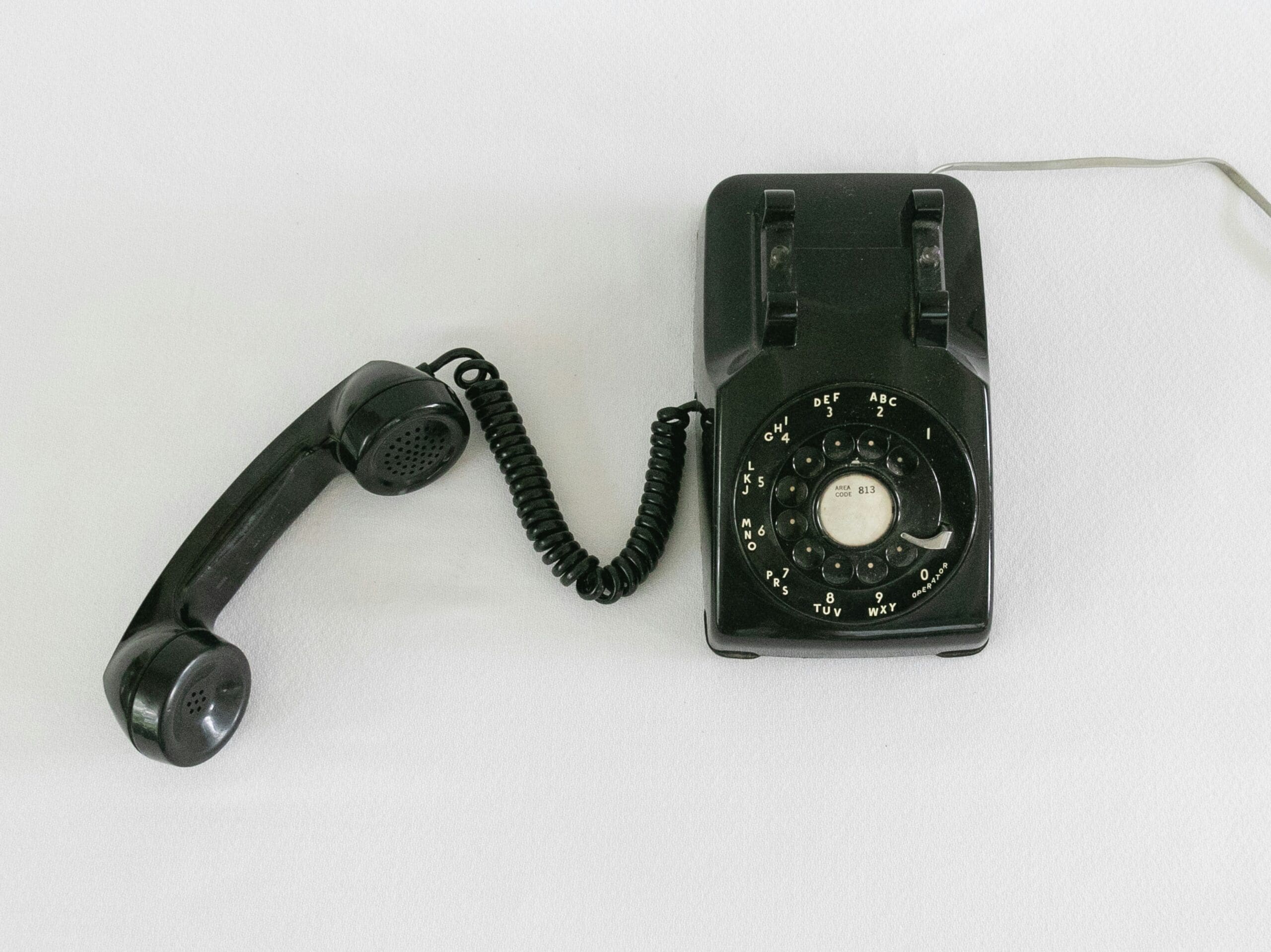 Black vintage rotary dial telephone with the handset off the hook, placed on a white surface.
