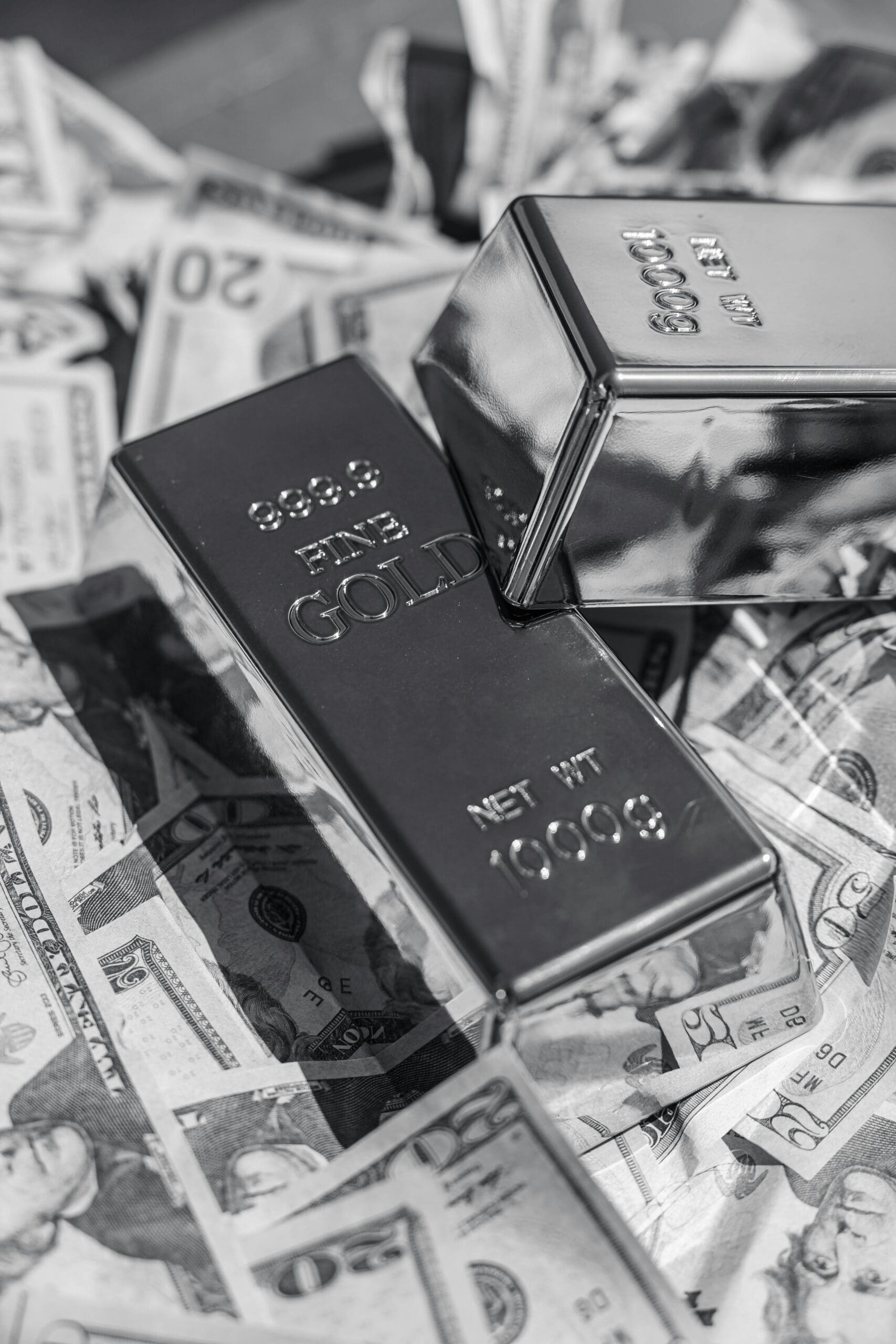 Two polished gold bars labeled "999.9 Fine Gold" rest atop a scattered pile of U.S. dollar bills in black and white.