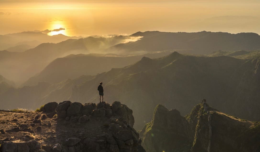 Person standing on rocky mountain peak at sunset, overlooking layered ridgelines and distant coastline under golden light.