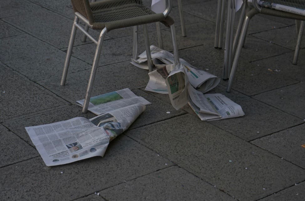 Crumpled newspapers scattered under café chairs on a stone-paved sidewalk.