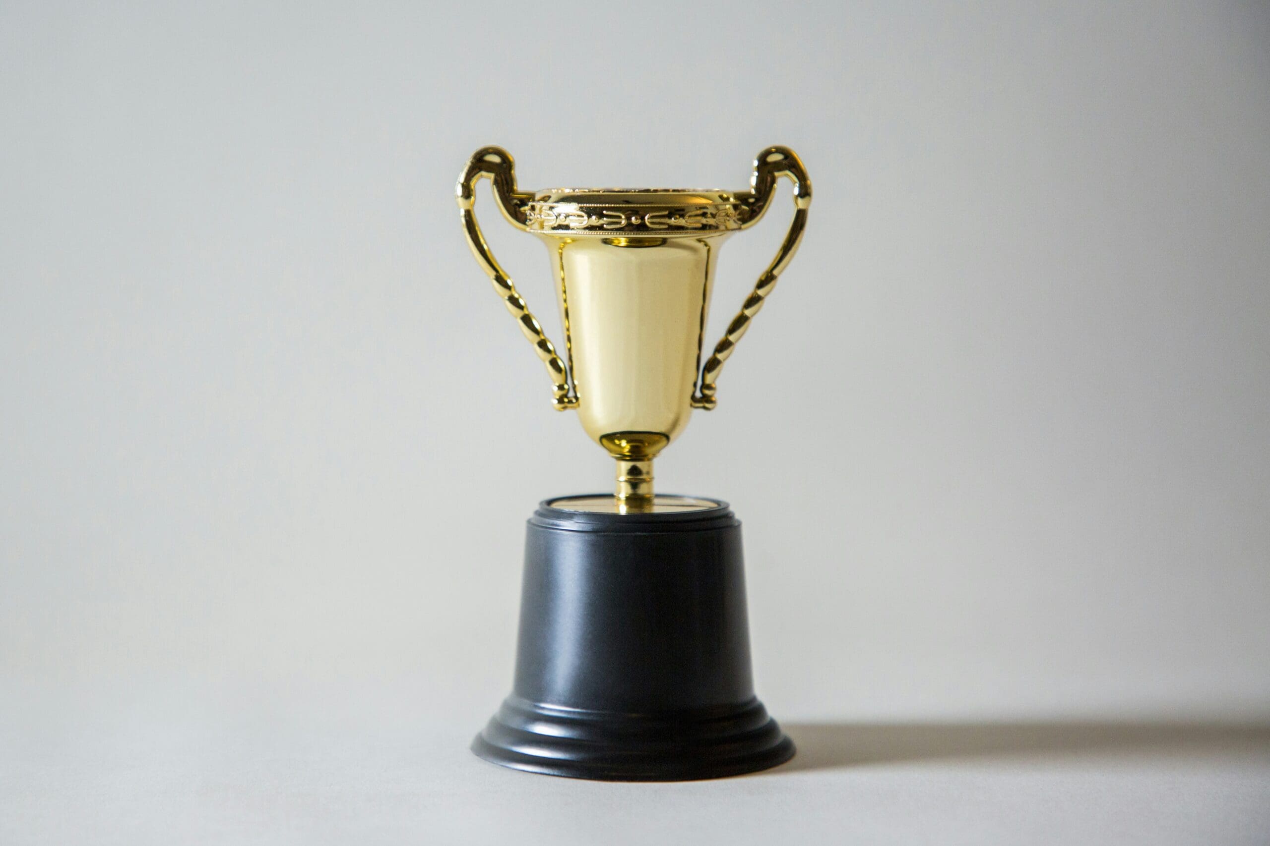 A small gold trophy with ornate handles on a black base, photographed against a plain light-gray background.