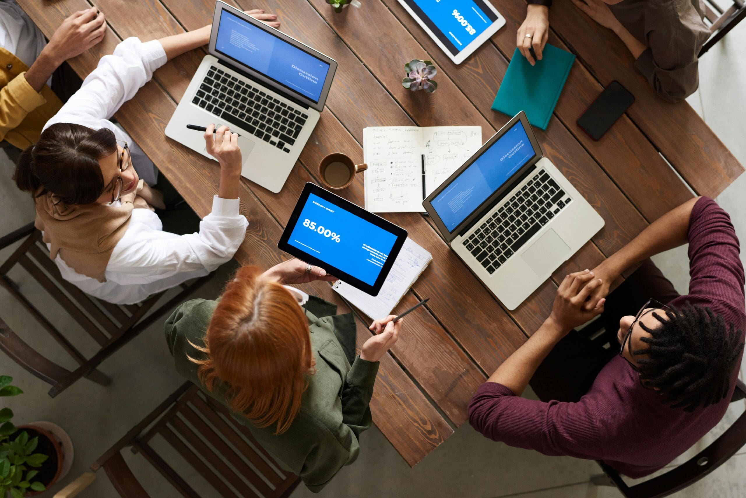 Overhead view of a diverse team working collaboratively around a wooden table with laptops, notebooks, and a tablet displaying data.