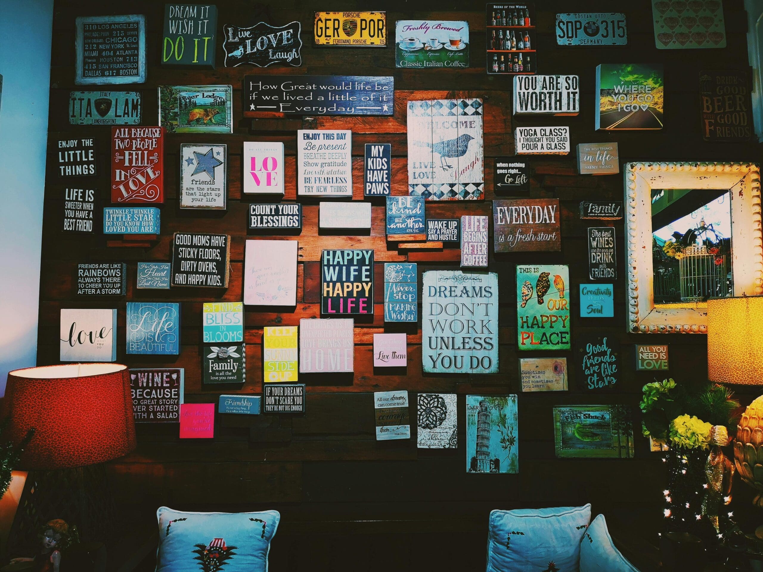 A warmly lit wall filled with vintage-style signs and quotes about love, life, and happiness, arranged above two armchairs with lamps and flowers.