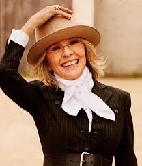 Smiling woman wearing a tan hat, glasses, black pinstripe suit, and white scarf, exuding confidence and timeless style.