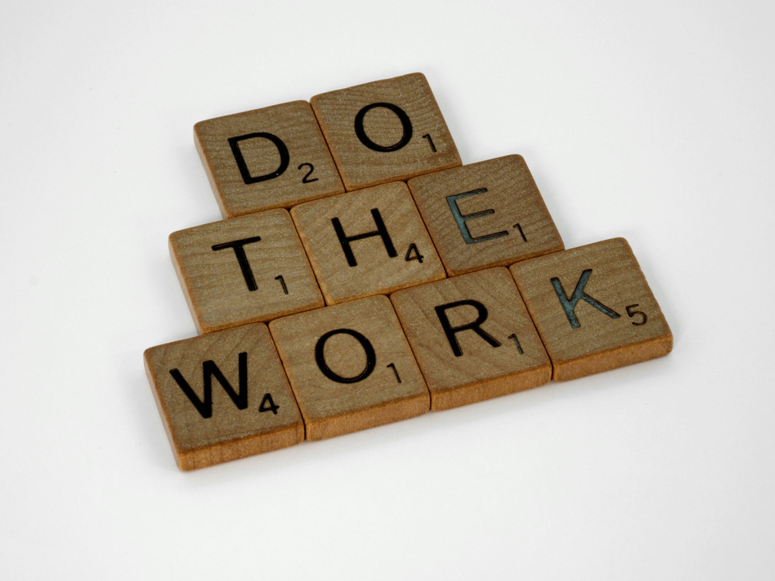 Wooden Scrabble tiles arranged to spell "Do The Work" on a white background.