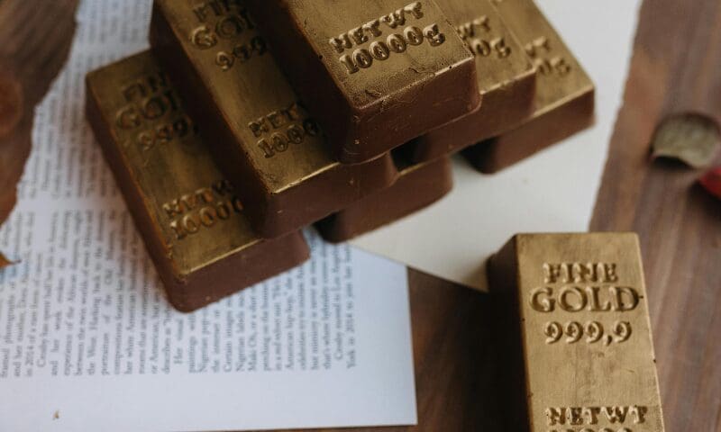 Close-up photograph of stacked gold bars with a warm, reflective surface. The lighting highlights their metallic texture and engraved markings, creating a luxurious and polished composition symbolizing wealth and financial value.