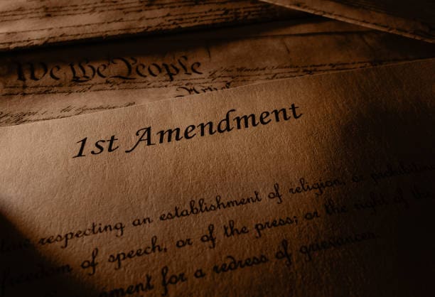 A sepia-toned close-up of a historical document showing the words "1st Amendment" with cursive script underneath, partially revealing the U.S. Constitution's opening phrase "We the People."