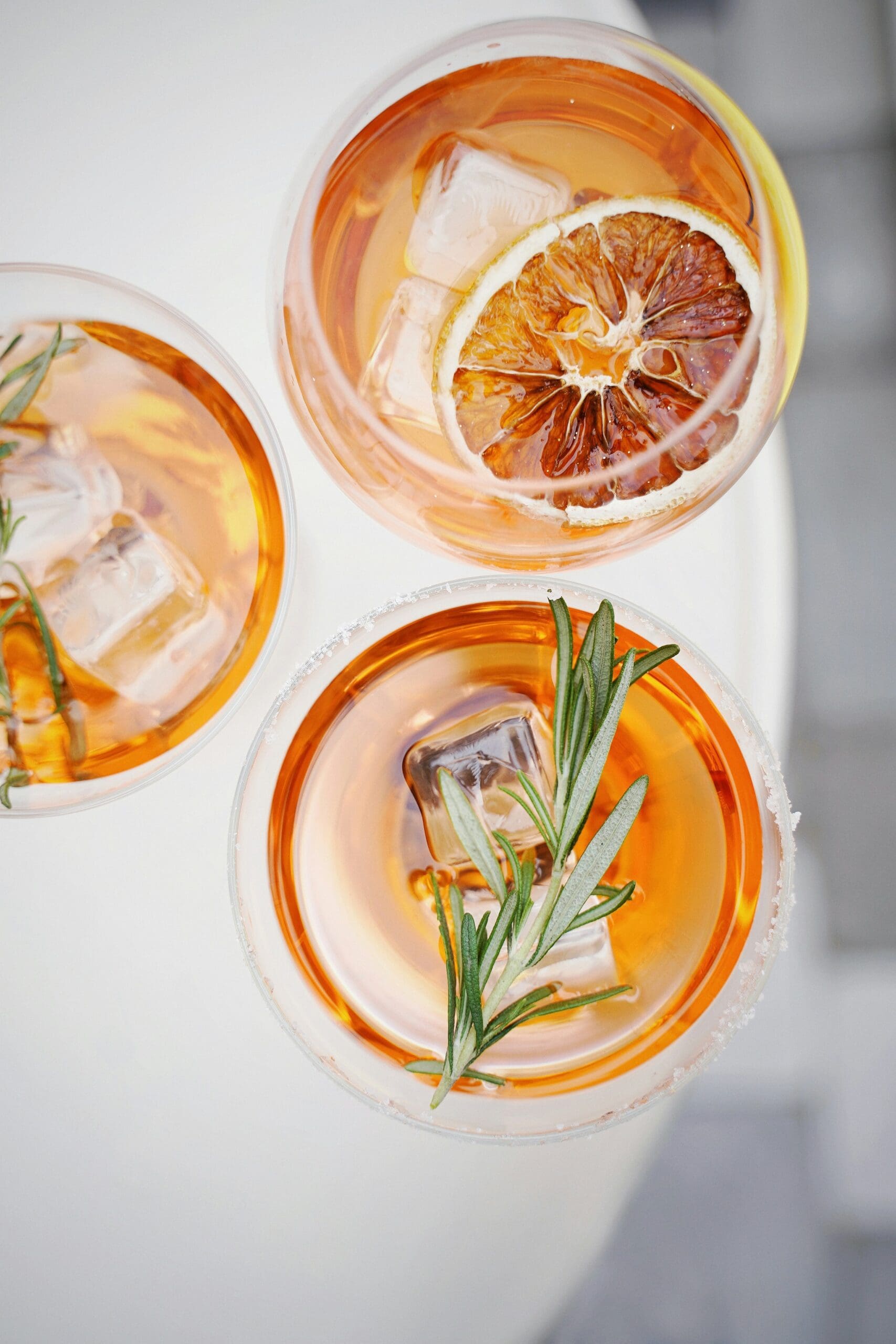 Overhead view of three golden cocktails with ice, rosemary sprigs, and a dried orange slice on a white table.