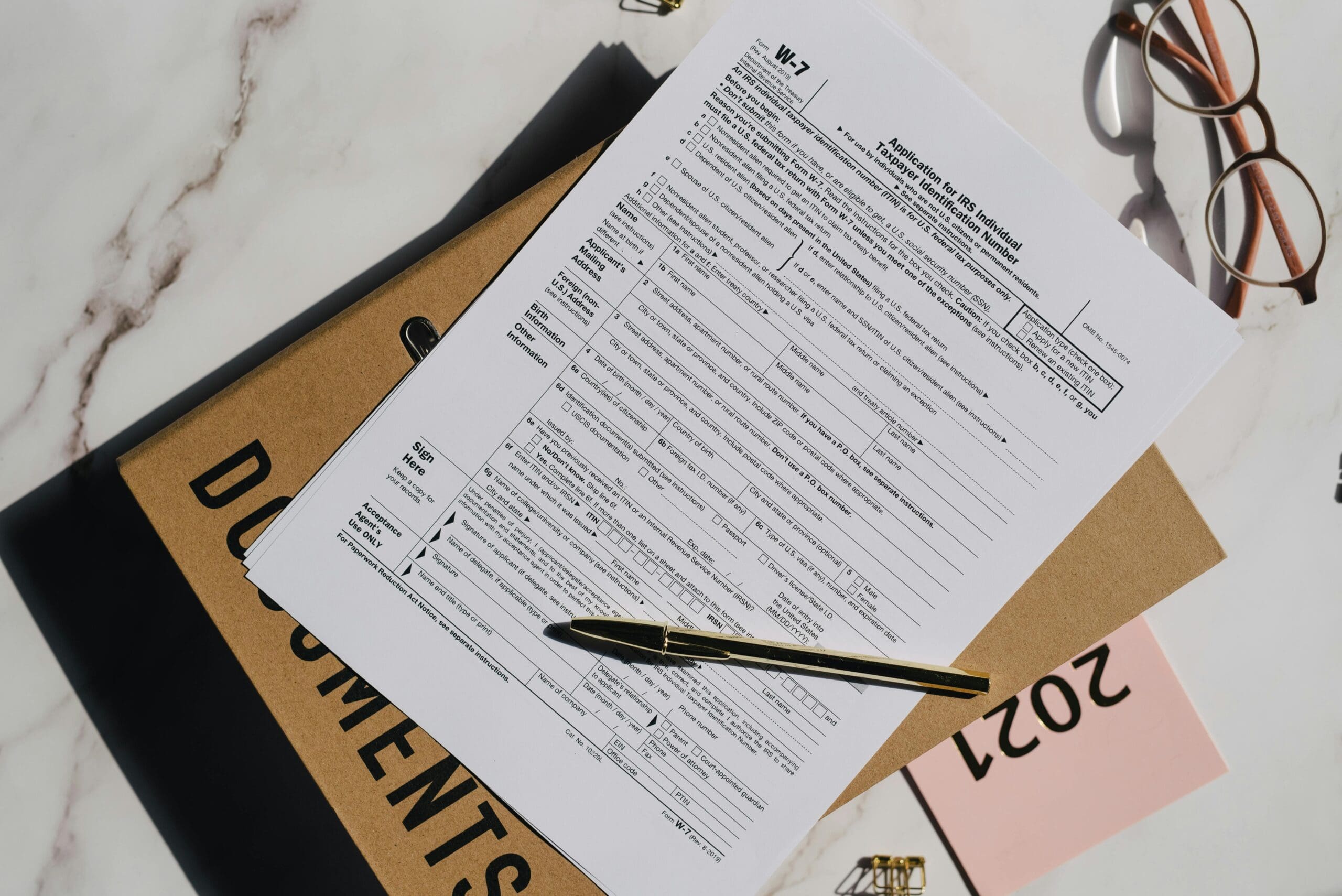 IRS W-7 tax form with pen, documents folder, and reading glasses on a marble desk.