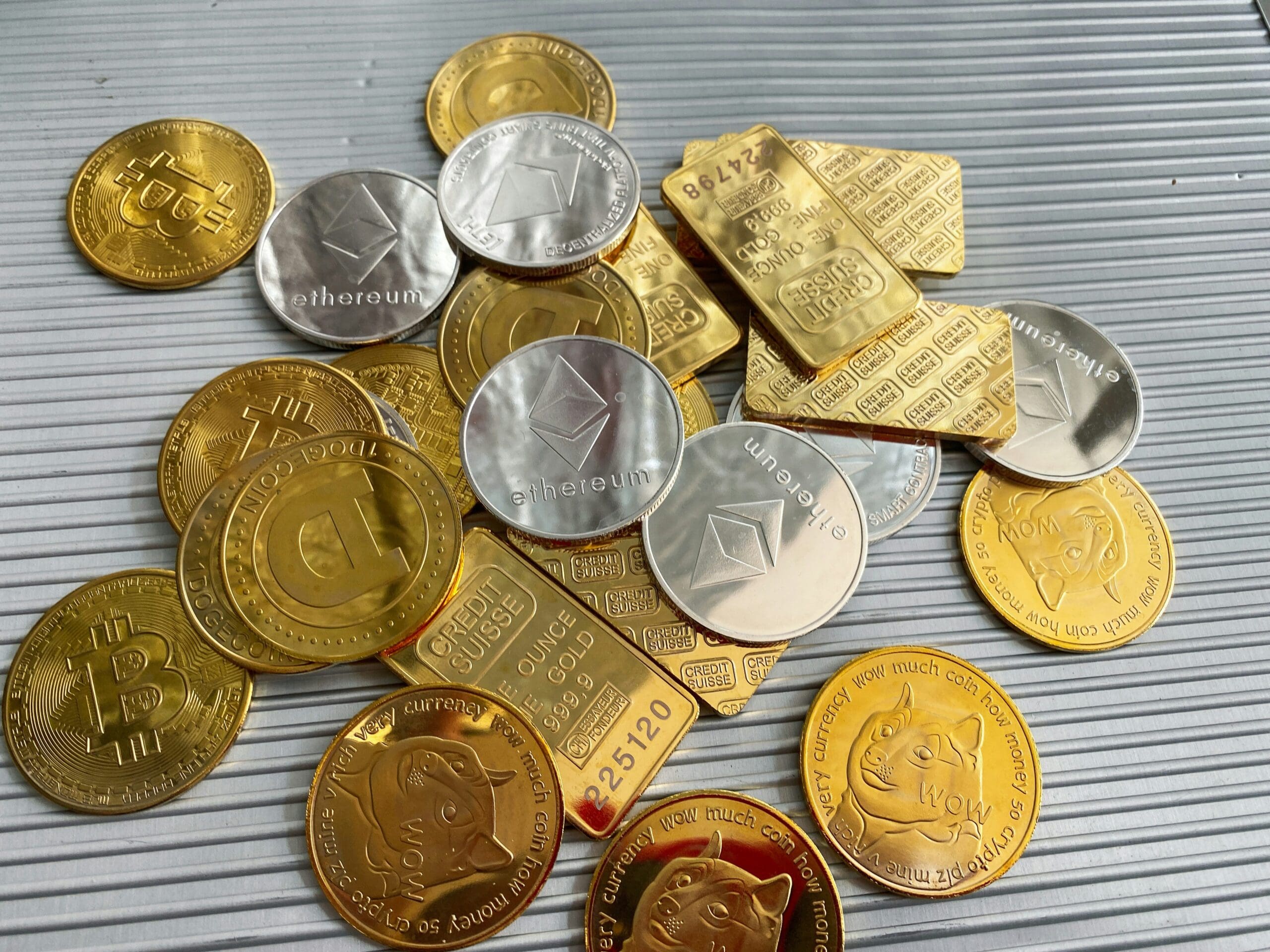 Assorted physical representations of cryptocurrencies (Bitcoin, Ethereum, Dogecoin) and gold bars scattered on a ribbed metallic surface.