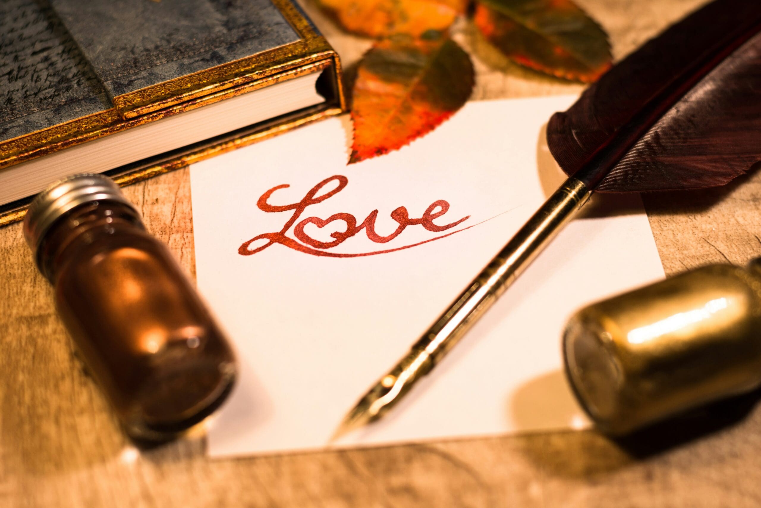 A handwritten “Love” on parchment beside a quill pen, ink bottle, and an antique book, all set on a wooden table with autumn leaves.