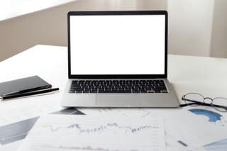 Open laptop with a blank white screen on a desk surrounded by financial charts, papers, a notebook, and eyeglasses, symbolizing data analysis, market research, or remote work in a professional setting.