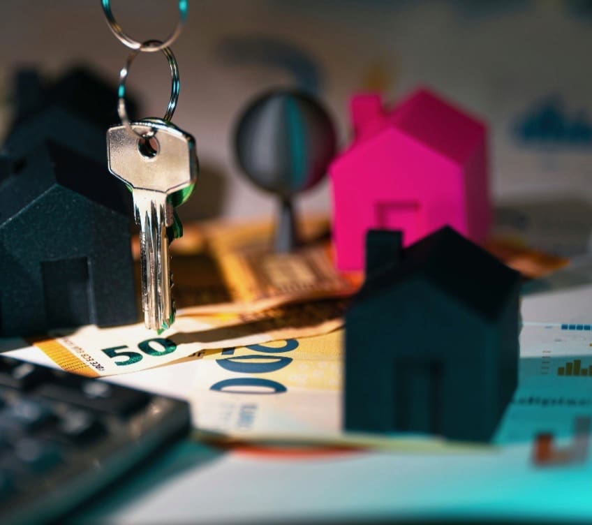 A close-up of house keys, small model houses, and euro banknotes representing real estate prices and housing affordability.
