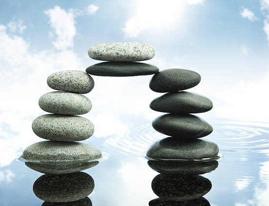 Stacked stones forming a balanced arch over calm reflective water with a bright sky and clouds in the background.