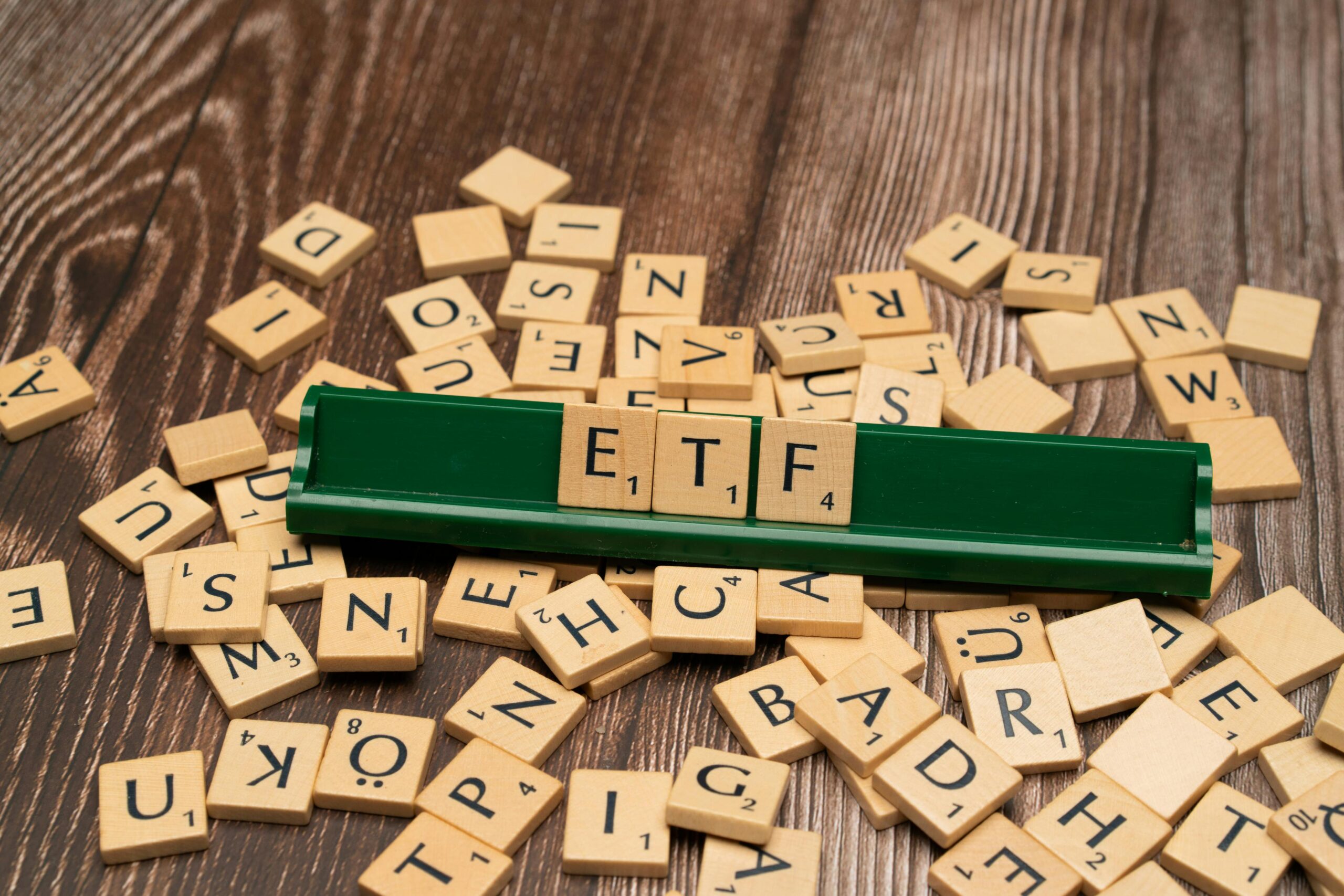 Wooden Scrabble-style letter tiles spelling “ETF” on a green tile holder surrounded by scattered letters on a wooden surface.