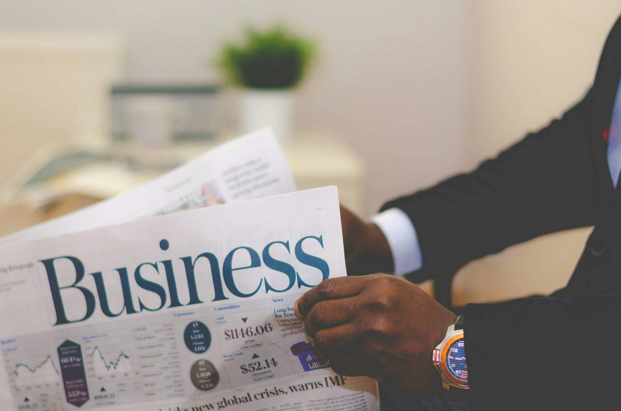 A person in a suit holding and reading a business newspaper with financial charts and market data visible.
