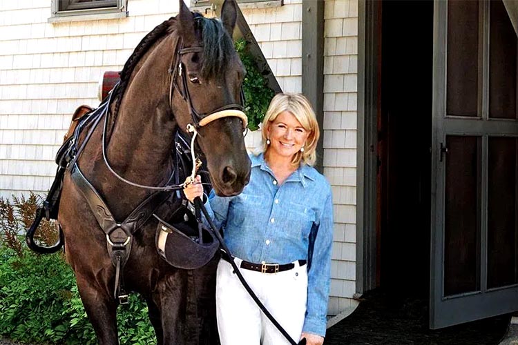 A woman stands beside a dark horse outside a barn, smiling as she holds the reins.