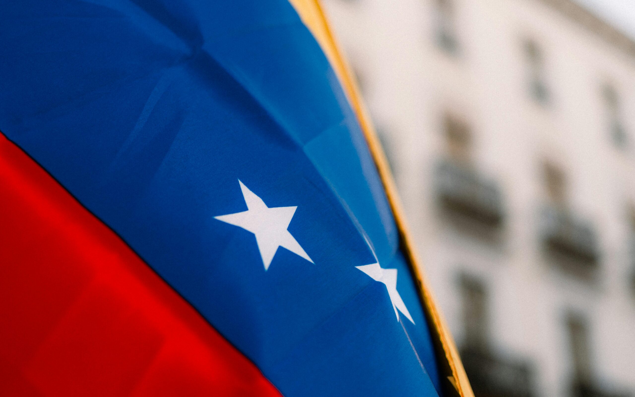 Close-up of the Venezuelan flag showing blue, red, and white stars with a blurred city backdrop.