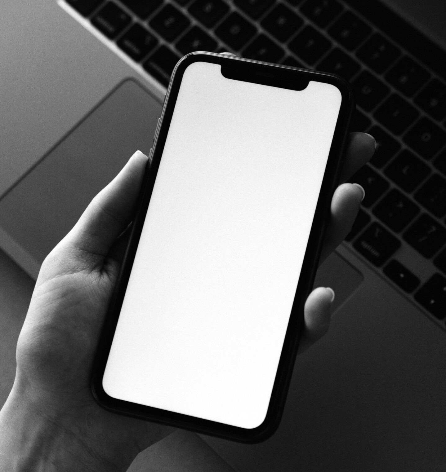 A hand holding a smartphone with a blank white screen, positioned above a laptop keyboard in a minimal black-and-white setting.