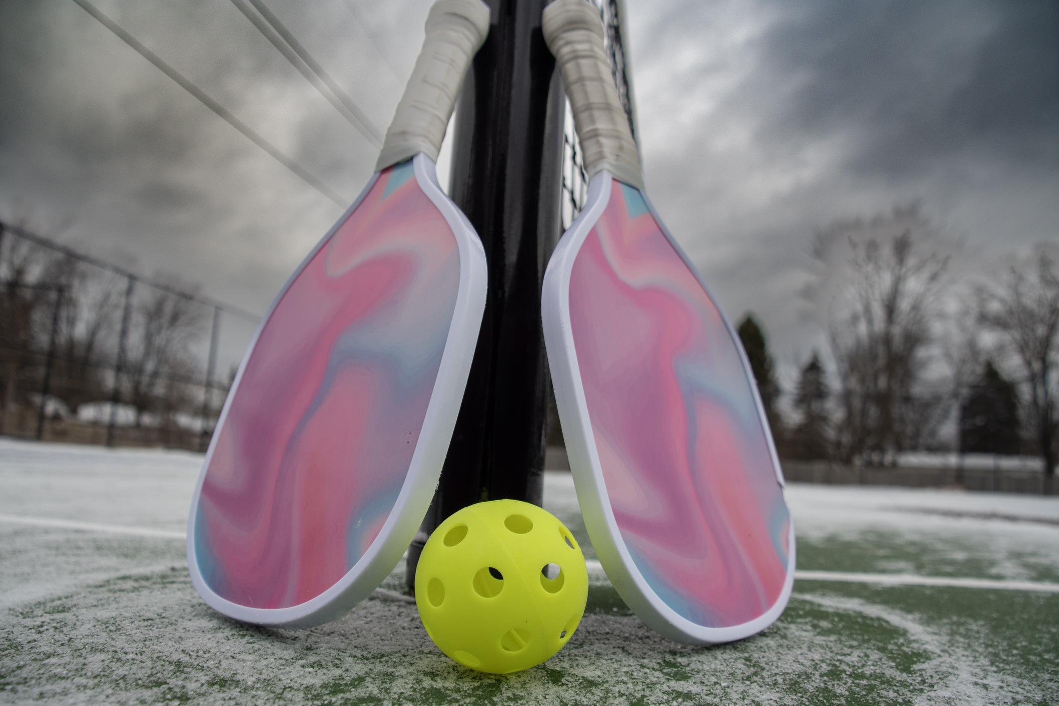 Two pastel pickleball paddles leaning against a net with a bright yellow ball on an outdoor court under a cloudy sky.