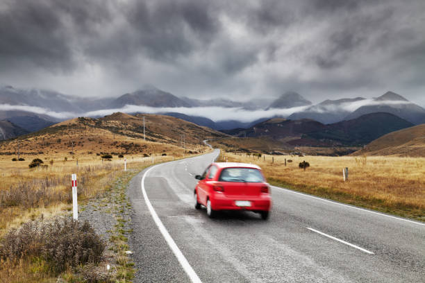 Red car driving along a winding rural road with mountains and dramatic storm clouds in the background.