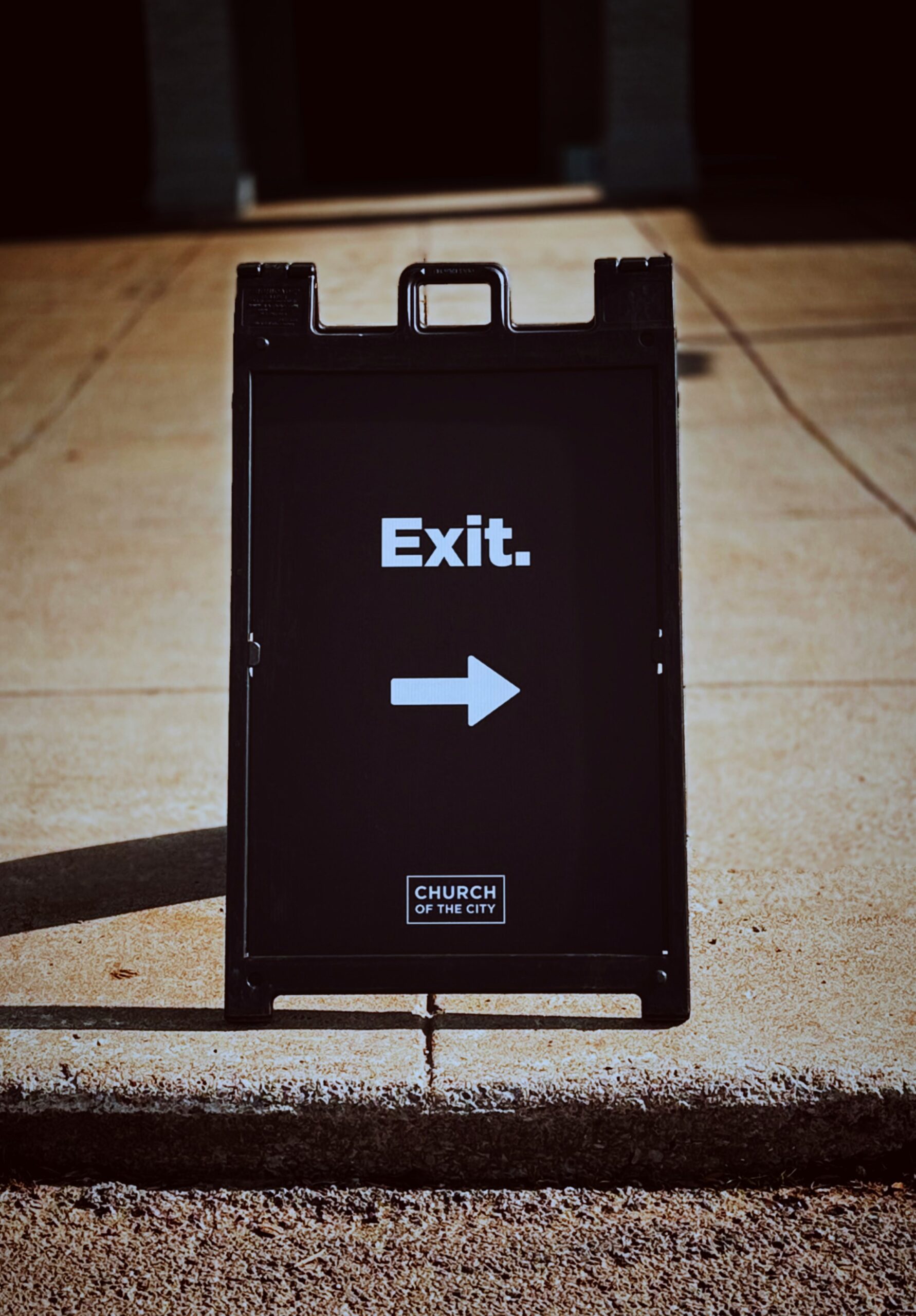 Black exit sign with a white arrow pointing right, placed on a concrete walkway in low light.