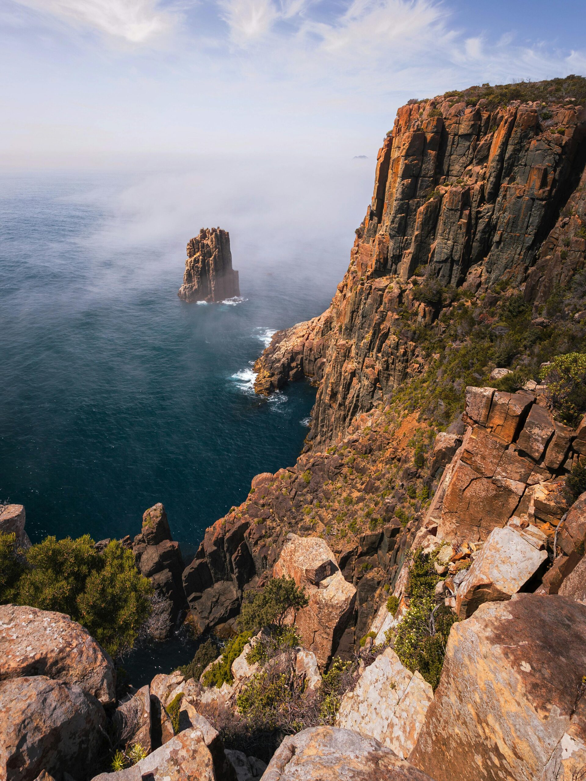 Rocky coastal cliffs overlooking the ocean with a tall sea stack rising from the water and light mist in the distance.