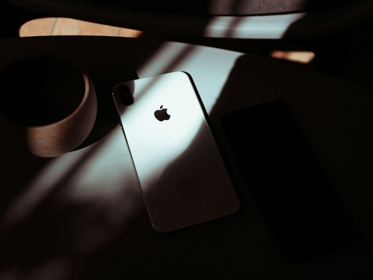 Backlit iPhone on a table beside a ceramic mug and another phone in shadow